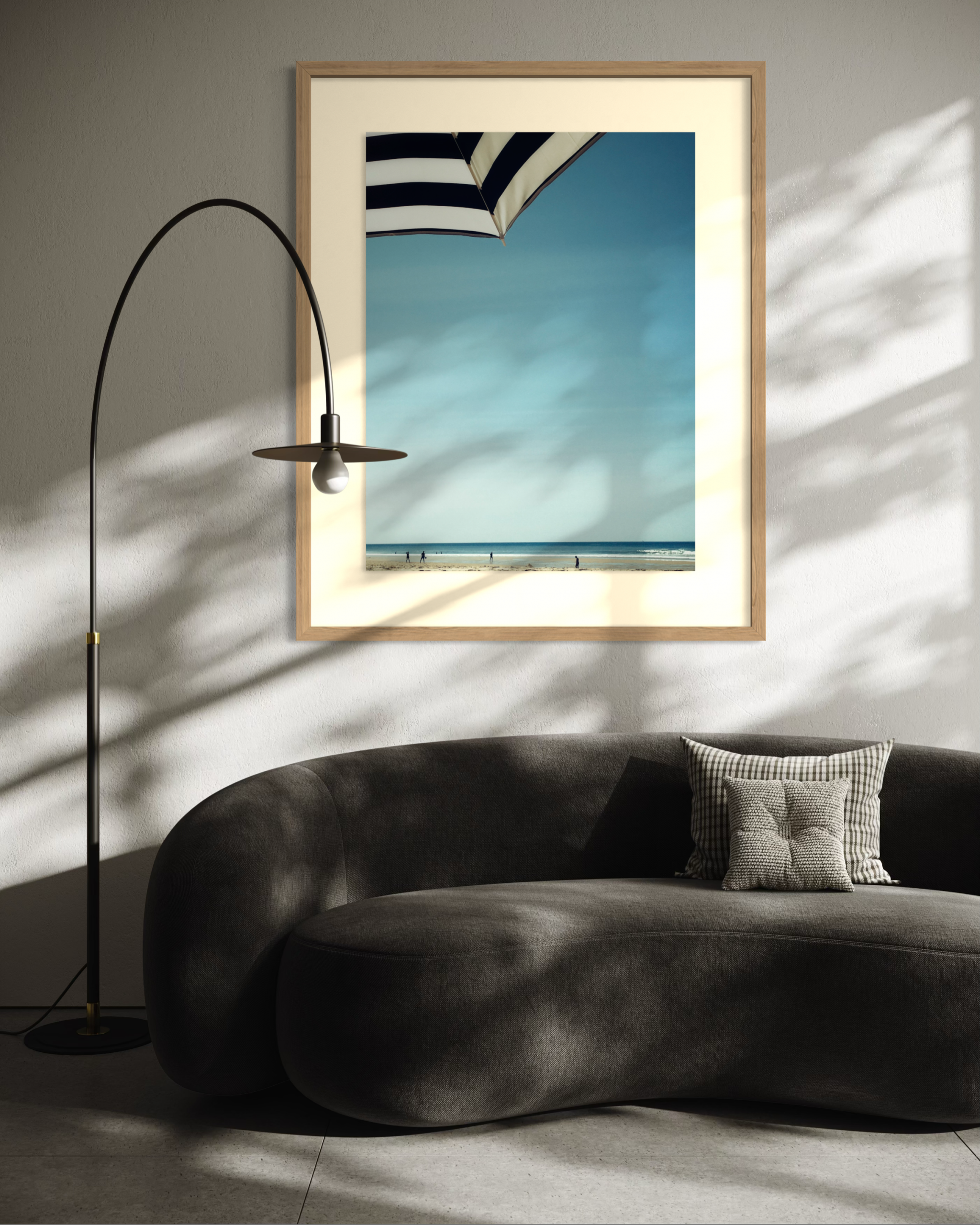 Salon moderne avec canapé arrondi gris, lampe arquée noire et tableau encadré représentant une plage sous parasol rayé noir et blanc