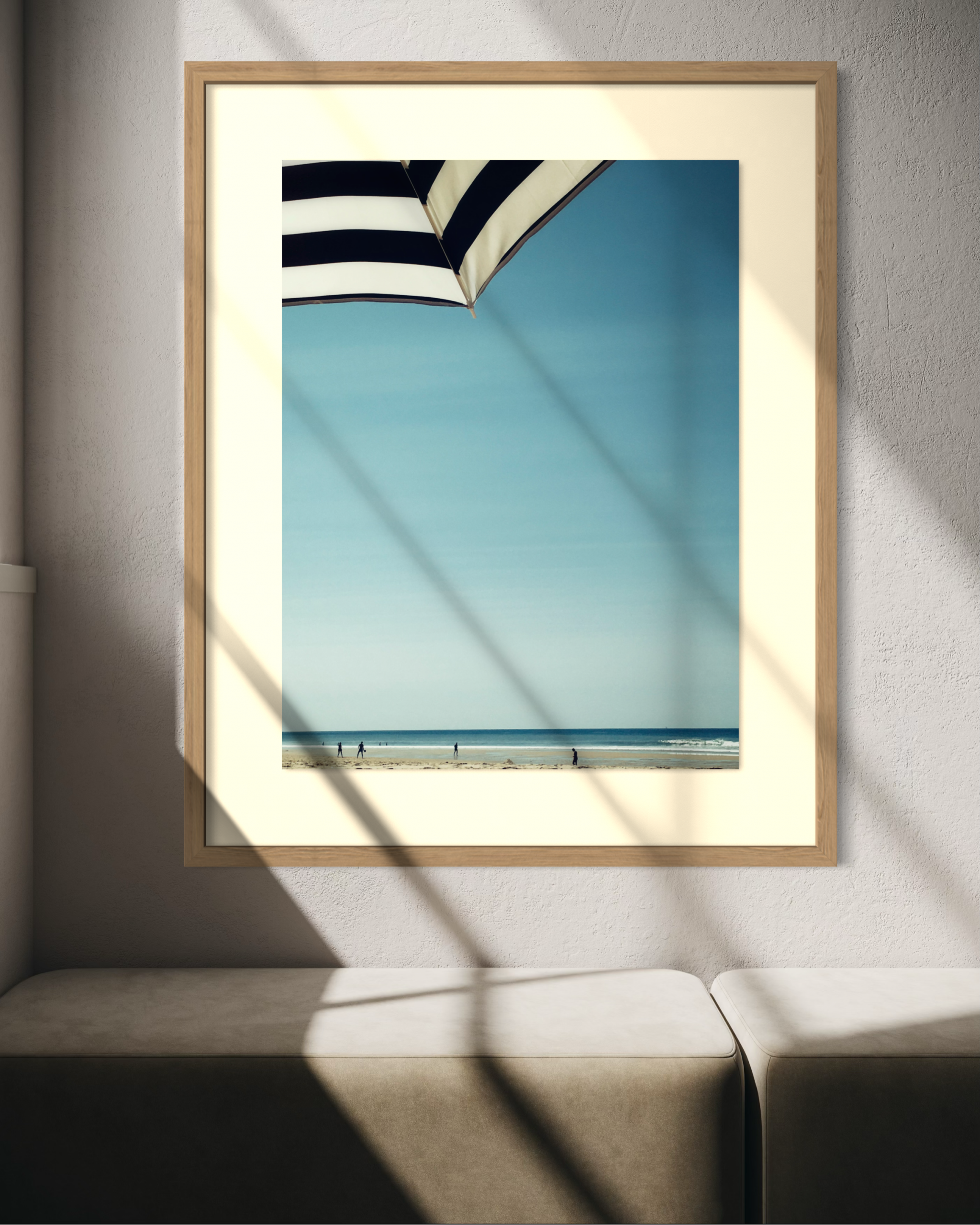Photographie encadrée d'une plage vue sous un parasol rayé noir et blanc, avec ciel bleu et silhouettes au bord de l'eau