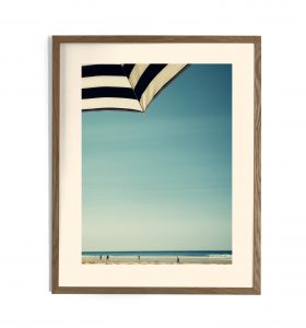 Impression encadrée d'une plage avec parasol rayé noir et blanc, ciel bleu et silhouettes de baigneurs au loin
