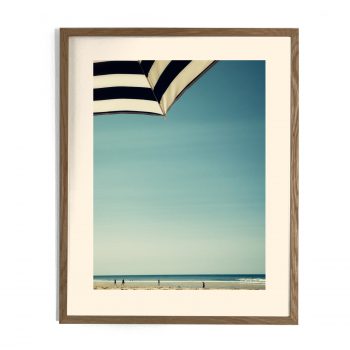 Impression encadrée d'une plage avec parasol rayé noir et blanc, ciel bleu et silhouettes de baigneurs au loin