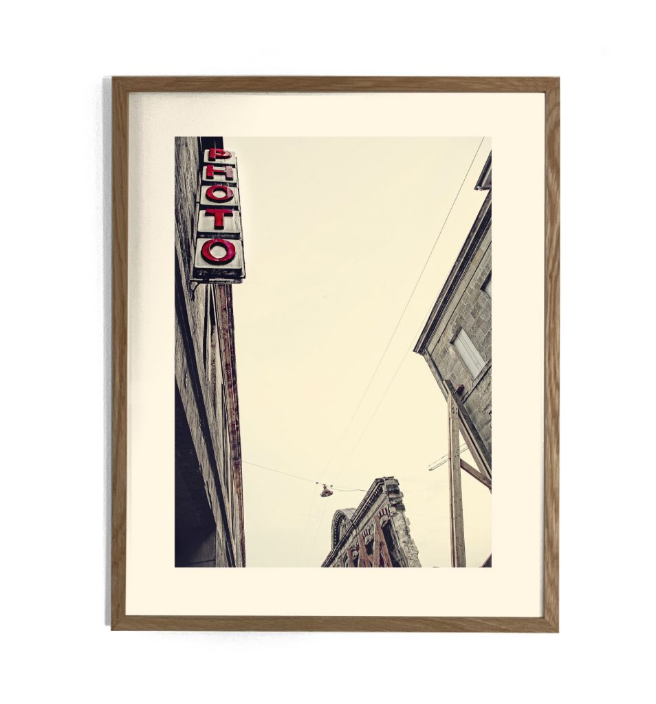 Photographie encadrée d'une enseigne verticale rouge « PHOTO » sur un bâtiment ancien, vue en contre-plongée urbaine
